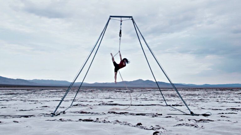 An image of a tall slanted structure made up of thin poles sits in a flat desert scene. An acrobat hangs from the center