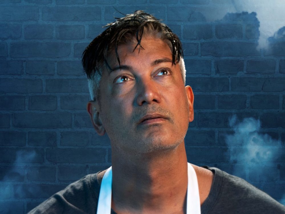 A man gazes upwards, behind him is a brick wall partially obscured by smoke