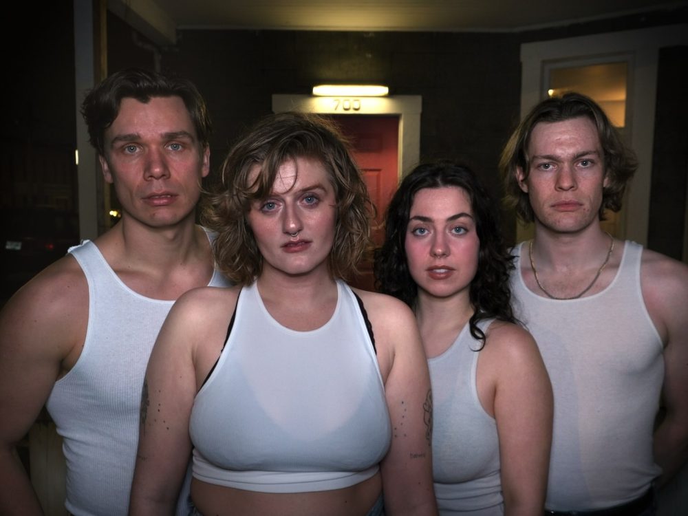 Two women and two men, all in white undershirts, stand close together in a dimly lit entryway.