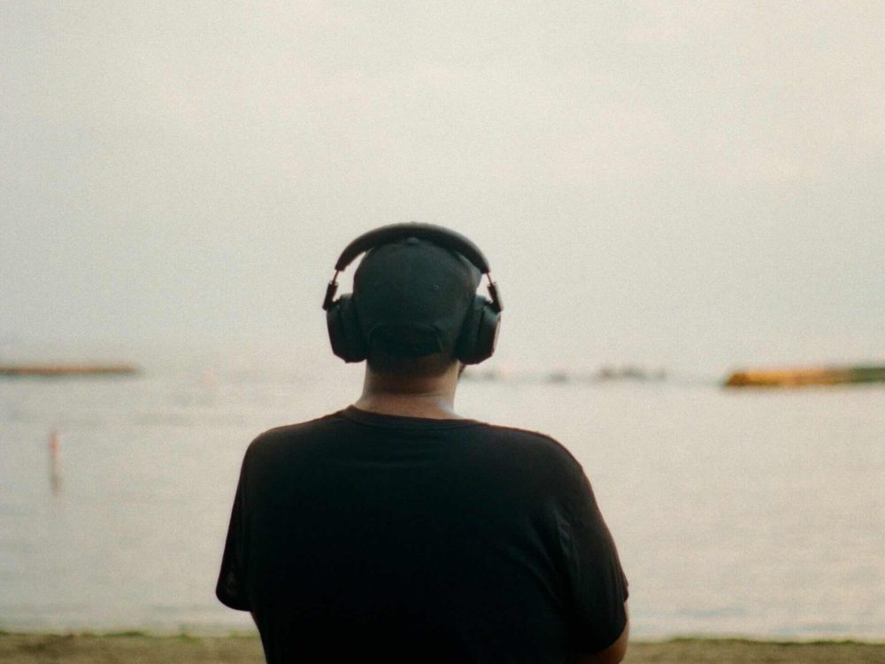 A person wearing a black t-shirt, black baseball cap and large headphones stands on a beach facing away towards a large body of water and a bright cloudy sky.