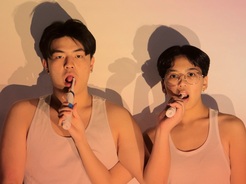 Two asian people with short hair brushing each others' teeth with electric toothbrushes. They are wearing white sleeveless undershirts.